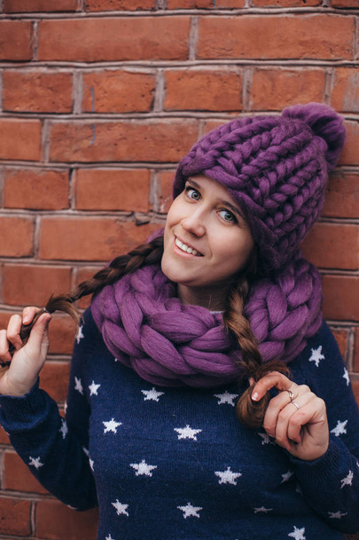 Brunette woman in purple knitted hat, knitted infinity scarf and blue sweater with white stars on it make posing with brick wall on the background - Photo, image