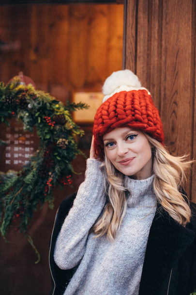Young blonde woman in red knitted hat with white pompon, grey sweater, black jacket and blue jeans make posing with Christmac cafe window on the background - Foto, afbeelding