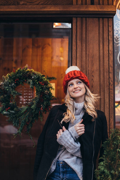 Young blonde woman in red knitted hat with white pompon, grey sweater, black jacket and blue jeans make posing with Christmac cafe window on the background - Fotoğraf, Görsel