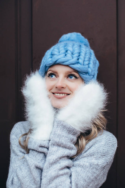 Young blonde woman in blue knitted hat, white fluffy mittens and grey sweater make posing with wooden door on the background - Fotoğraf, Görsel