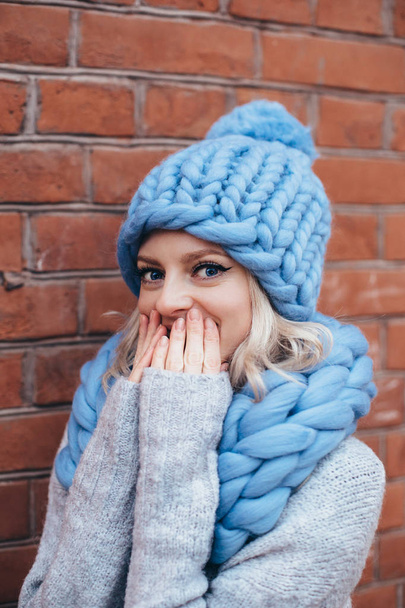 Young blonde woman in blue knitted hat, blue khitted infinity scarf and grey sweater make posing with brick wall on the background - Photo, image