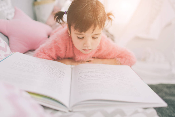 Cute little kid girl is reading a book at home. Funny lovely child having fun in kids room. - Foto, Imagem