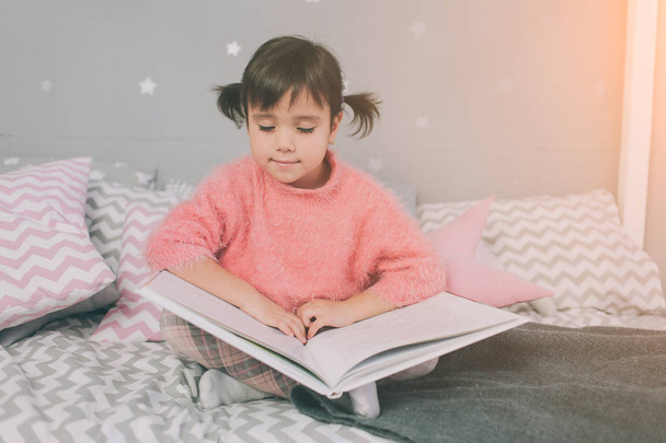 Cute little kid girl is reading a book at home. Funny lovely child having fun in kids room. - Foto, Imagem