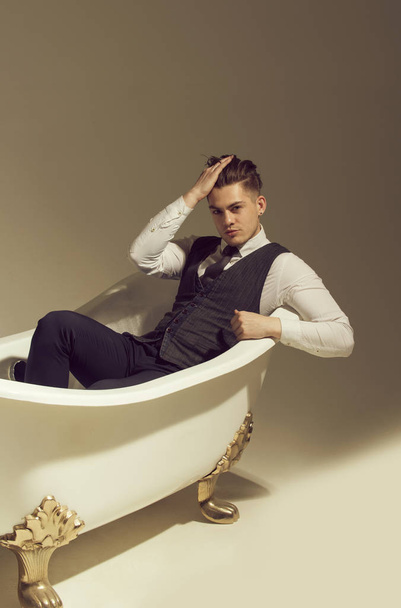 Handsome man or businessman with stylish haircut, hair, in fashion business wear, vest, tie, shirt and pants relaxing in classic bath, white tub, on grey background - Photo, image