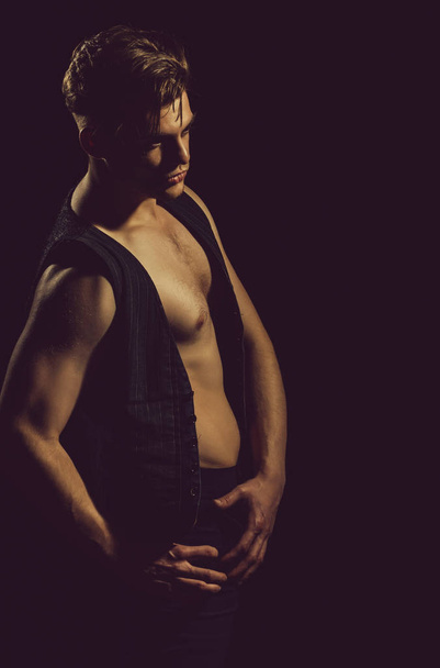 Handsome man or young caucasian macho, athlete, bodybuilder, with stylish haircut, hair, posing in unbutton vest with muscular torso, hands, arms, with biceps, triceps on black background - Fotó, kép