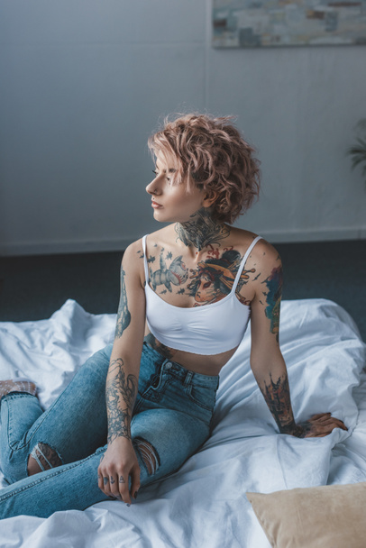 attractive trendy tattooed girl with pink hair sitting on bed - Foto, imagen