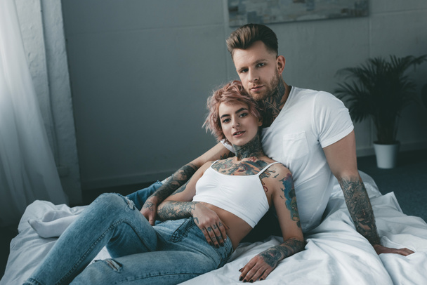 tattooed couple looking at camera and relaxing on bed - 写真・画像