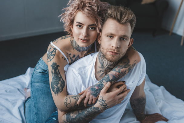 tattooed girlfriend looking at camera and hugging her boyfriend on bed in bedroom - Фото, изображение