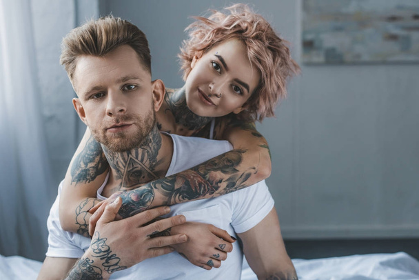 smiling tattooed girl hugging her boyfriend on bed in bedroom - Photo, Image