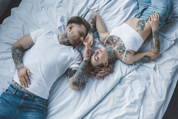 top view of tender tattooed lovers holding hands and relaxing on white bed - Foto, Imagen