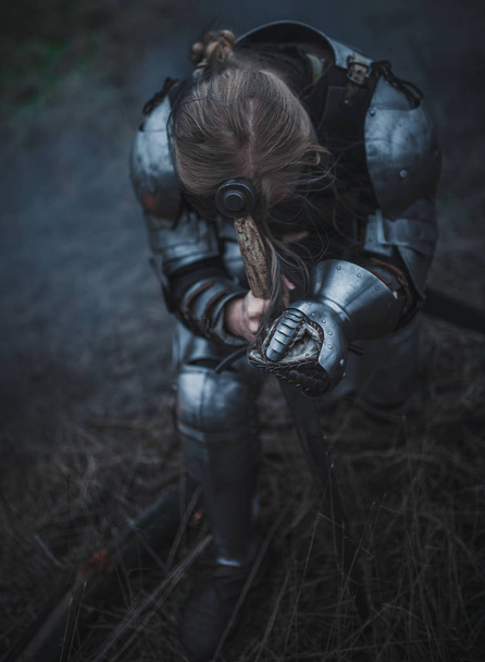 Menina na imagem de Jeanne d 'Arc em armadura e com a espada em suas mãos ajoelha-se contra o fundo da grama seca
. - Foto, Imagem
