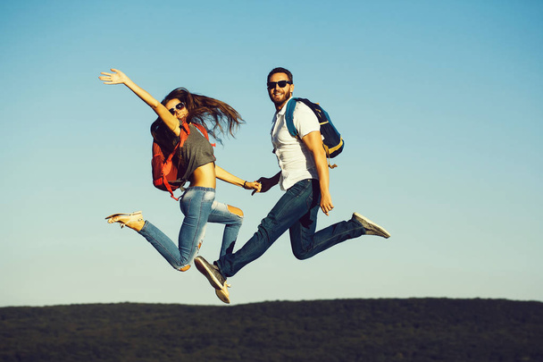 jumping happy couple of handsome bearded man in white shirt and pretty sexy cute woman with background with backpack outdoor on sunny day on blue sky background - Photo, image