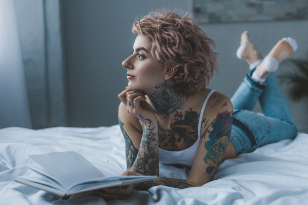 thoughtful tattooed girl lying on bed with book - Photo, image