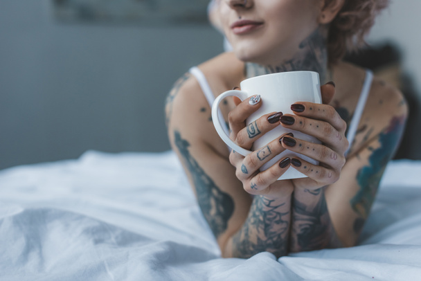 cropped view of tattooed girl drinking coffee in bed at morning  - Foto, Imagem