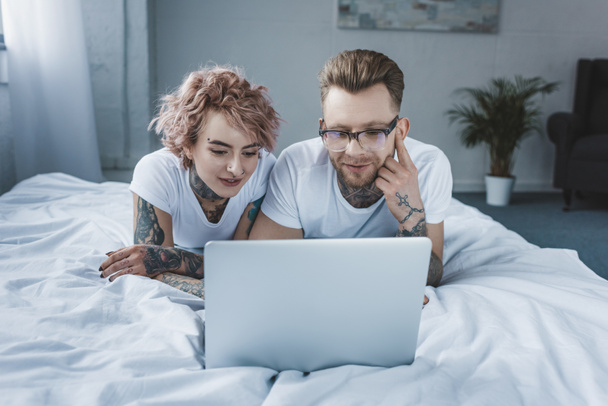 tattooed smiling couple using laptop together in bed - 写真・画像