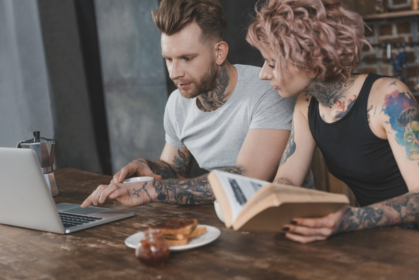tattooed couple using laptop and reading book during breakfast in kitchen - Фото, зображення