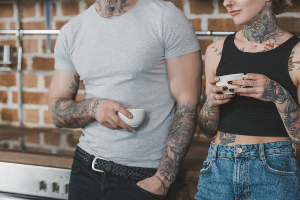 cropped view of tattooed couple drinking coffee in kitchen at morning - Foto, Imagen