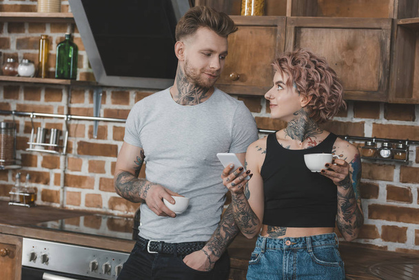 young tattooed couple drinking coffee and using smartphone in kitchen at morning - Фото, зображення