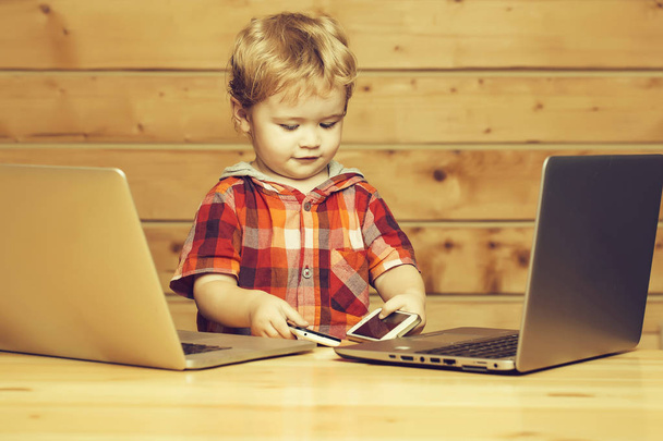 Cute baby boy child with blond curly hair plays on two phones and laptop computers on wooden background - Zdjęcie, obraz