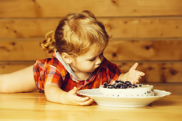 Cute baby boy child with blond curly hair eats delicious cake with blueberries lying on wooden table - Photo, image