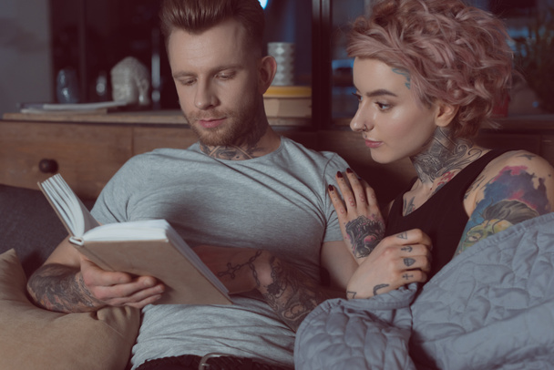 pareja tatuada descansando y leyendo libro en casa
 - Foto, Imagen