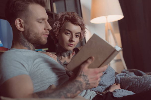tattooed couple reading book while lying on sofa at home - 写真・画像