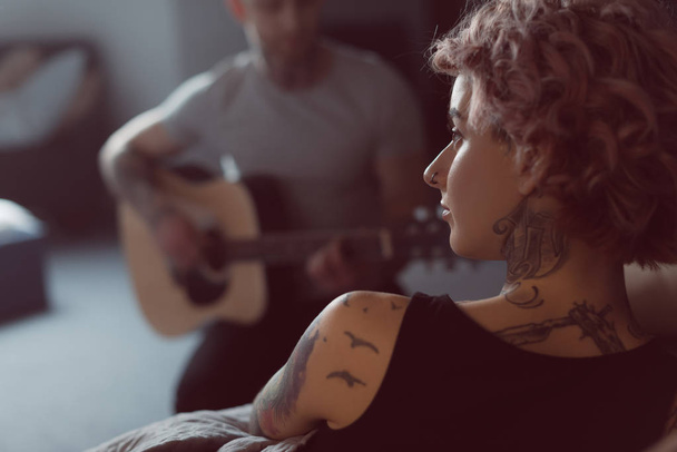 ο φίλος που παίζει για την ακουστική κιθάρα για τατουάζ φίλη του στο σπίτι - Φωτογραφία, εικόνα