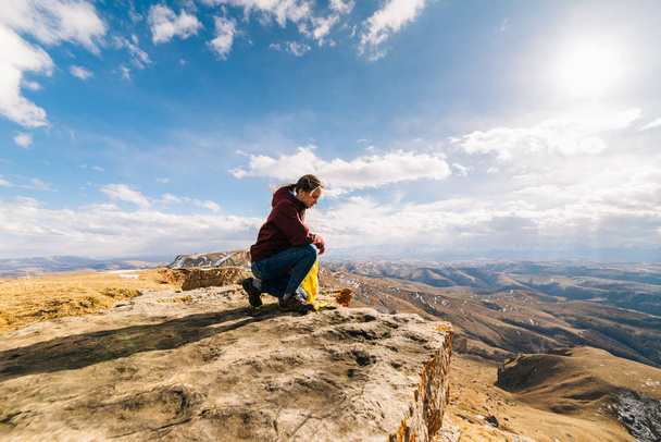 active young girl sits at the edge of the mountain, enjoys nature and the sun, holds a yellow backpack - 写真・画像