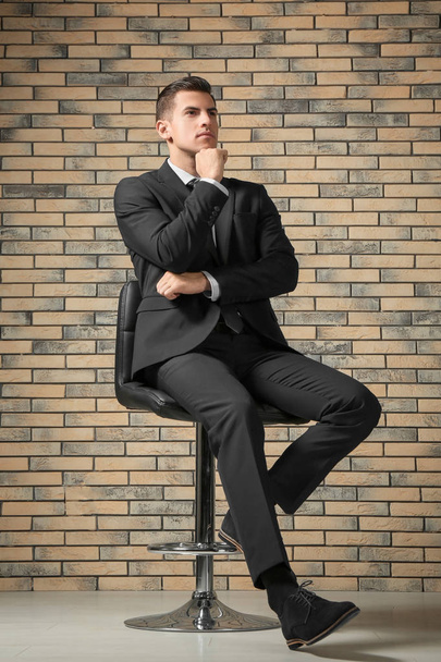 Handsome man in elegant suit sitting on chair near brick wall - Photo, image