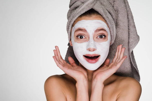 funny young girl with a towel on her head applied a white moisturizing face mask - Fotó, kép