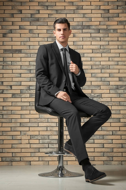 Handsome man in elegant suit sitting on chair near brick wall - Photo, Image