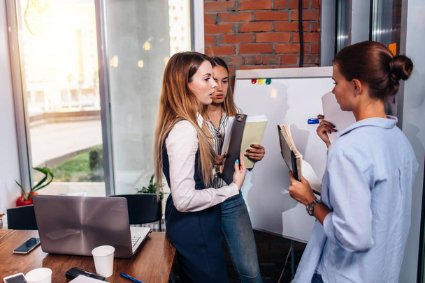 Три молодые бизнесвумен делятся новыми бизнес-идеями, держащие заметки, стоящие у доски в офисе
 - Фото, изображение