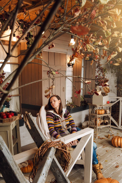 Beautiful young happy brown-haired woman, dressed in a sweater, jeans and yellow shoes, sitting on the porch in chair in autumn time. Courtyard, lit by flashlights, with dry fall leaves and pumpkins. - Фото, изображение