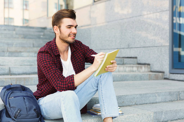 Νεαρός φοιτητής κάνοντας σημειώσεις που κάθονται στα σκαλοπάτια - Φωτογραφία, εικόνα