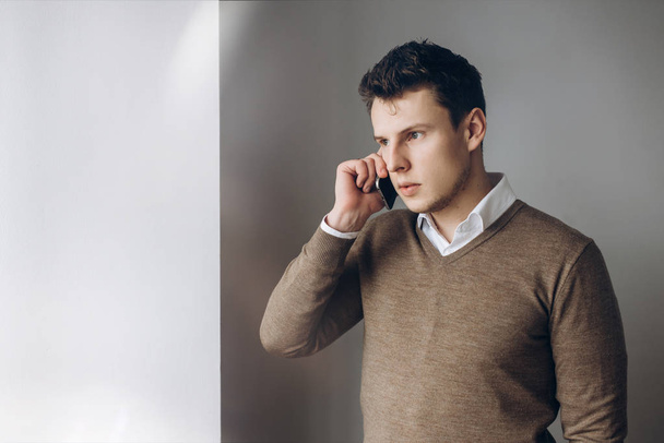 Young handsome guy with a serious face speaks on a smartphone and looks out the window - Fotografie, Obrázek