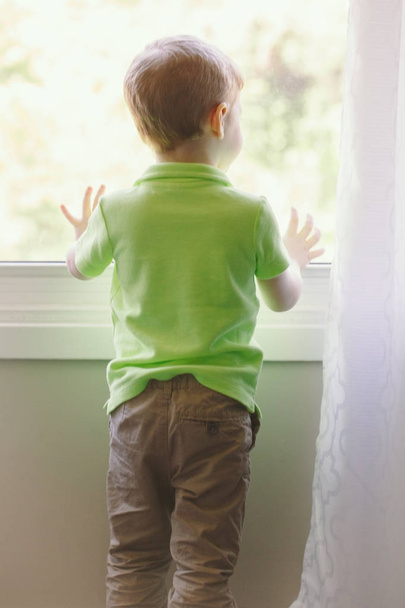 Portrait of white Caucasian child boy at home looking out of window outside. View from back. Kid waiting for someone - Foto, imagen