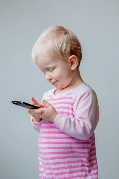 Cute adorable white Caucasian blond baby with blue eyes making a call, playing with mobile cell phone with funny expression on her face, lifestyle new generation technology - 写真・画像