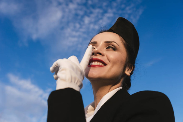 happy smiling female stewardess in uniform puts finger to lips, under blue sky - Foto, imagen