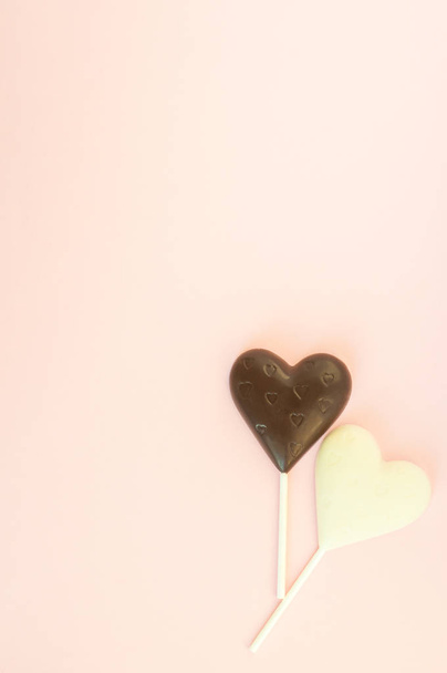 Black and white chocolate candy in the shape of a heart on a stick on a light pink background. Vertical Card to the Valentine's Day. - Fotó, kép
