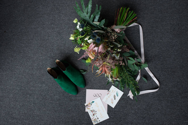 Green bridal shoes, rich green wedding bouquet with pink ribbons and a wedding complimentary lying on a grey floor  - Photo, image