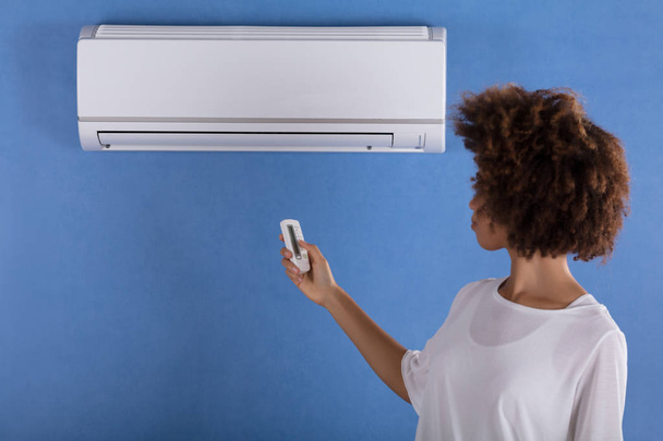 Young Woman Adjusting Air Conditioner Mounted On Blue Wall With Remote Control - Φωτογραφία, εικόνα