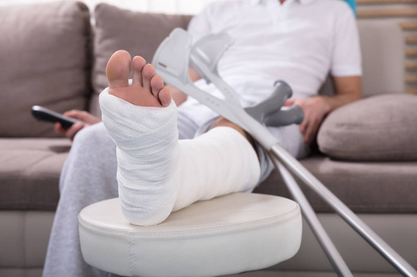 Young Man With Broken Leg Sitting On Sofa Holding Remote - 写真・画像