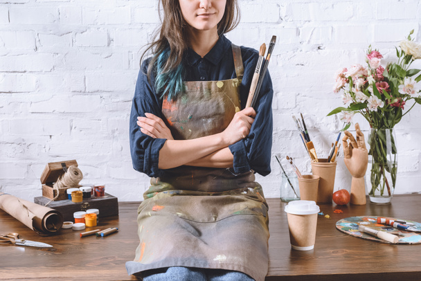 cropped image of female artist sitting on table with crossed arms and holding brushes  - Photo, image