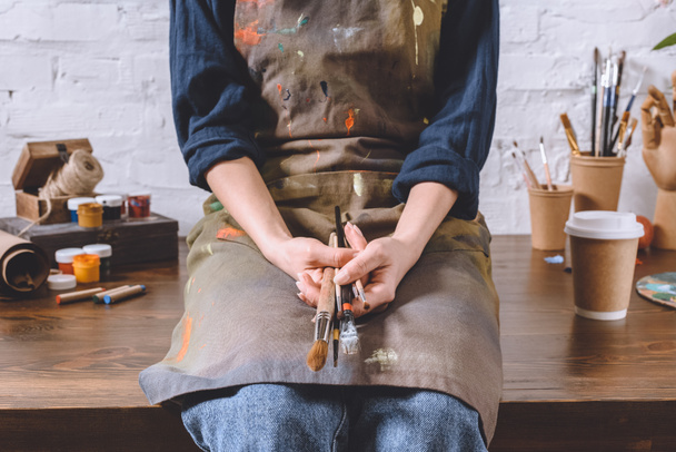 imagem cortada de artista feminina sentada na mesa e segurando escovas
  - Foto, Imagem