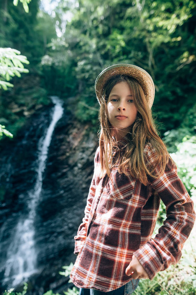 Schoolgirl in shirt and straw hat posing against waterfall background. Model in casual clothes outdoors facing camera. Vacations, relaxation, weekend, tourism, travel concept - Φωτογραφία, εικόνα