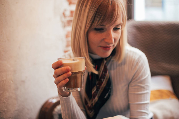 Young but mature independent woman, with neck scarf drink latte in see through cup while reads book or text on smartphone, short blond hair and feminine features. Coffe morning routine - Foto, imagen