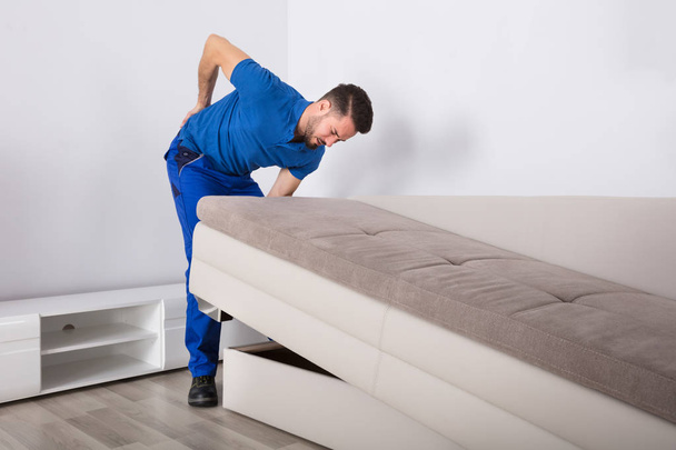 Young Male Mover Suffering From Backpain While Lifting Sofa In Living Room - Photo, Image