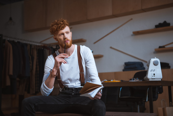 pensive young male fashion designer looking away while taking notes in notebook at workplace - 写真・画像
