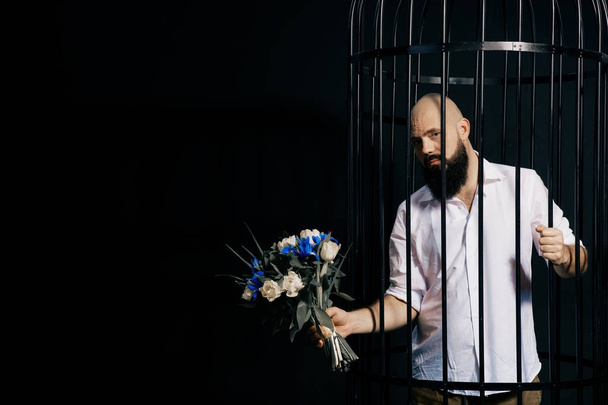 bearded man holding out a bouquet with flowers. The brutal man in the cage over black background - Фото, изображение