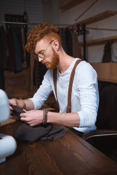 jonge mannelijke mode-ontwerper in brillen houden van stof tijdens workshop - Foto, afbeelding